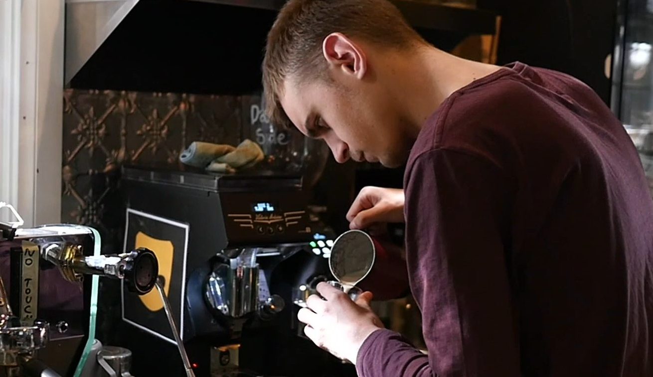 Young man pouring steamed milk from a jug into a cup. Standing in front of a coffee machine.