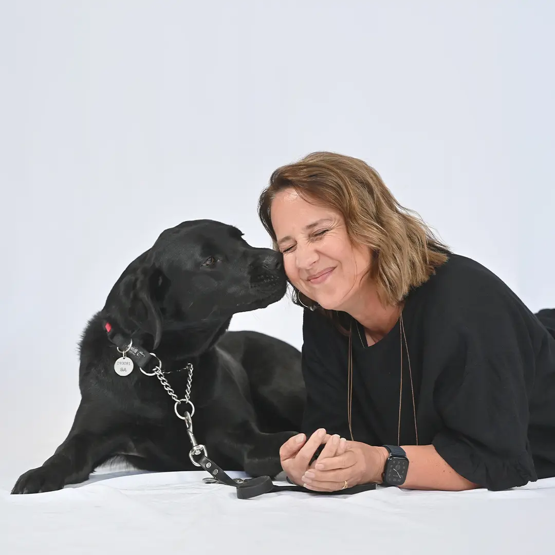 Lana a black guide dog lying down on the ground next to handler Lisa. Lana is touching her nose against Lisa's cheek
