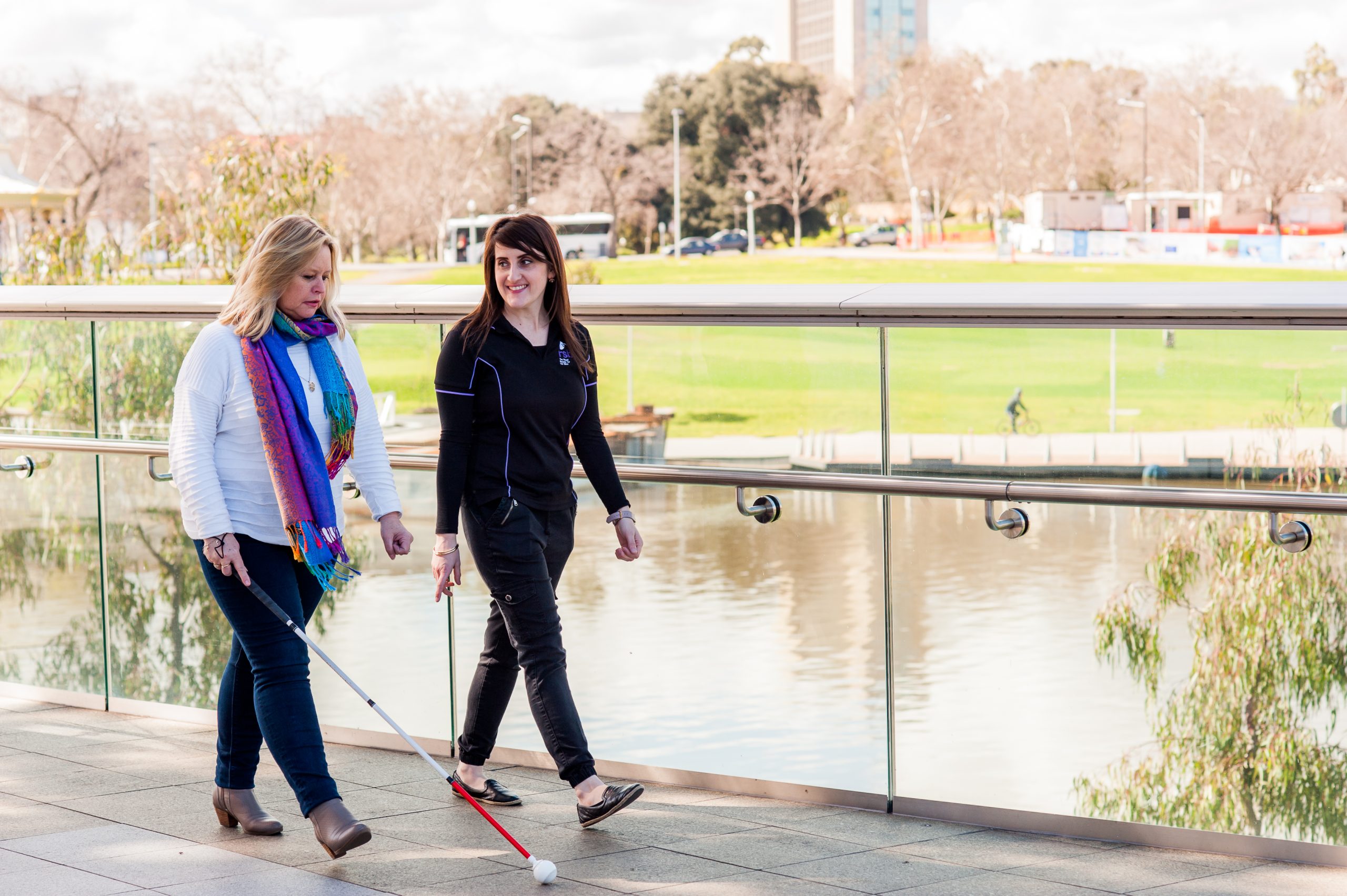 Two woman walking next to each other along the river Torrens. The lady on the left is holding a white cane. 