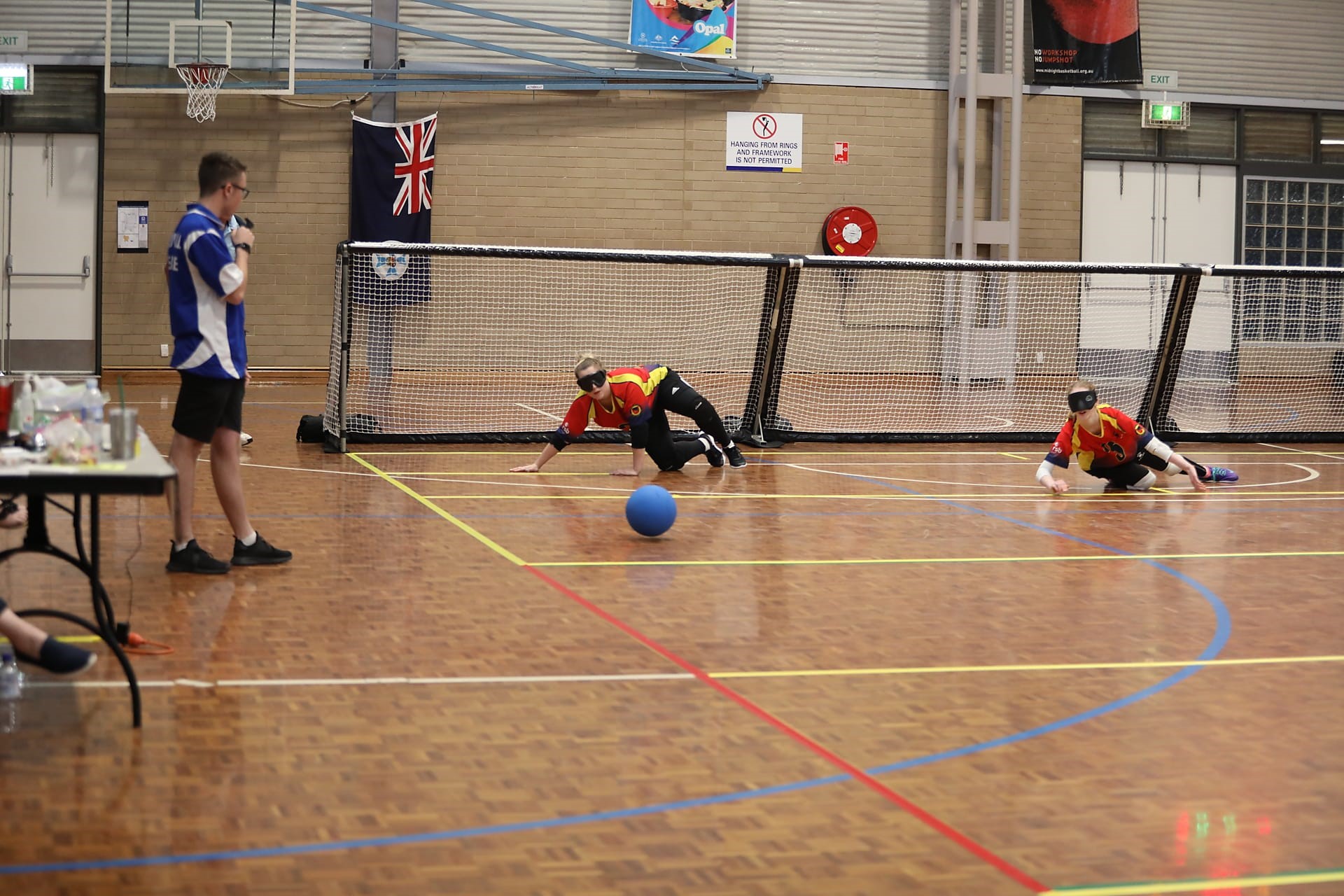 A blue ball rolls towards two women lying on the ground. A man on the left side of the court holds a microphone and is looking at the women. 