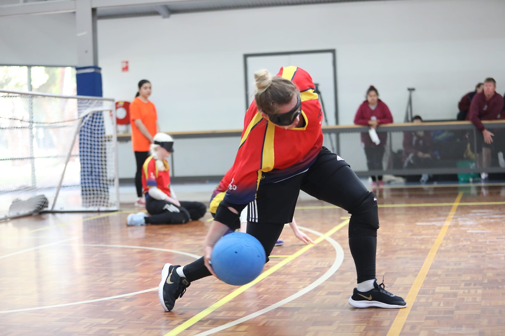 Nikita is wearing a red top and black pants. Her hair is in a bun and she is wearing black glasses. She has stepped her left leg forward and her right leg back. She is holding a blue ball in her right hand. 