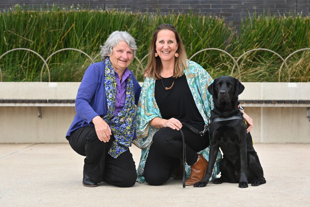Two women kneeling on the ground next to a black Guide Dog.